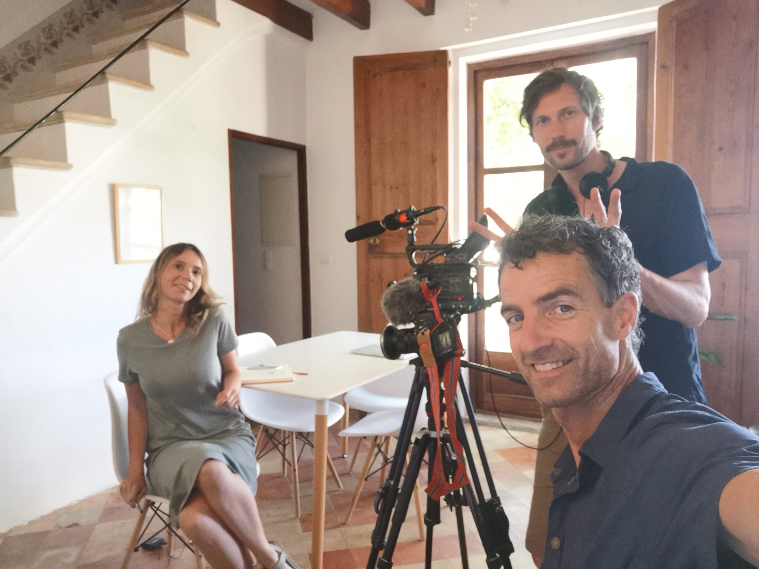 Three people in their thirties smiling into the camera. A professional camera is between them.