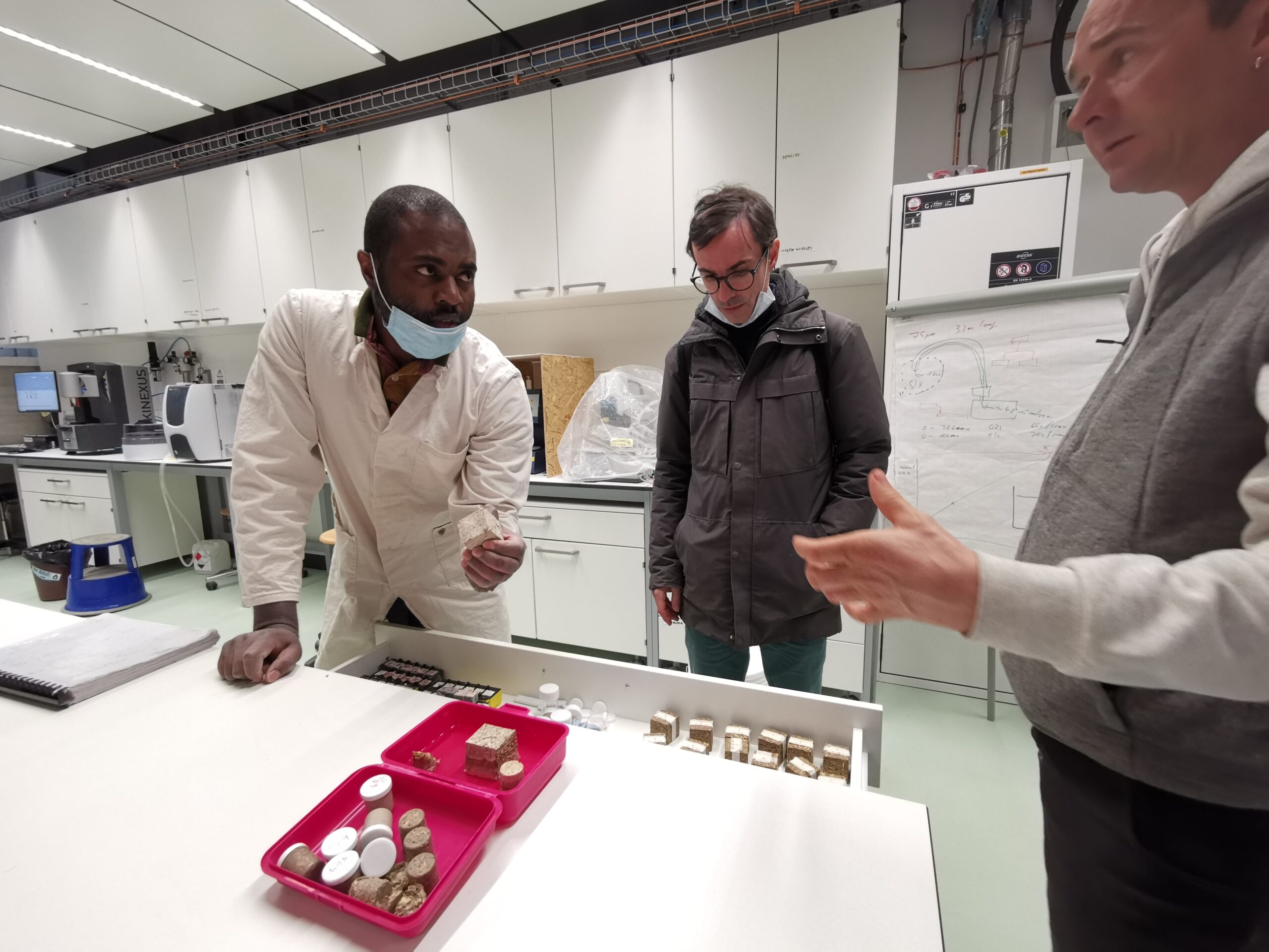 Three people in a lab. A black person in a lab coat is showing the two other several samples.